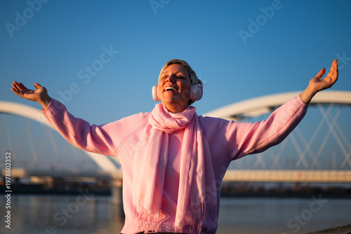 Portrait of happy woman with arms outstretched enjoying music with wireless headphones while sitting by the city river and smiling on a sunny day.