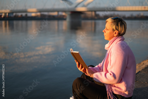 Portrait of urban woman with short blonde hair reading a book while enjoying sitting by the city river on a sunny day.