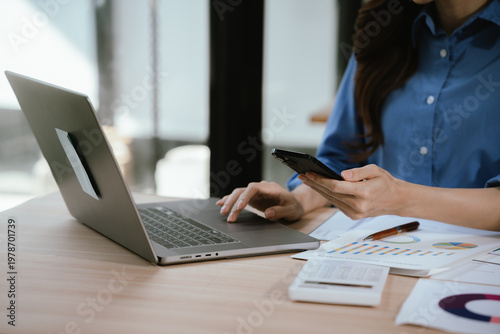 Close-up of hands typing on a laptop at a wooden desk with charts and a small flower pot, in a bright workspace with natural light from a window.