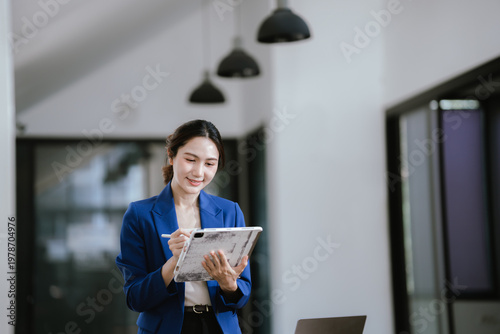 A professional businesswoman standing in a modern office, using a tablet and stylus, with a laptop on the desk behind her.
