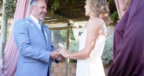 Couple holding hands pink-drape pergola groom speaking vows gowned bride listening Indian officiant