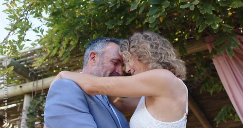 Couple pressing foreheads, holding each other under pergola, sharing affection in white lace dress