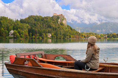 Schilderij op canvas A young woman sits in a rented wooden rowboat on Lake Bled, with the medieval Bled Castle in the distance at early autumn in the Julian Alps, at Bled Slovenia