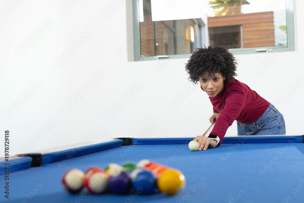 Fototapeta premium African American woman wearing burgundy top and jeans aiming cue at home pool table, copy space