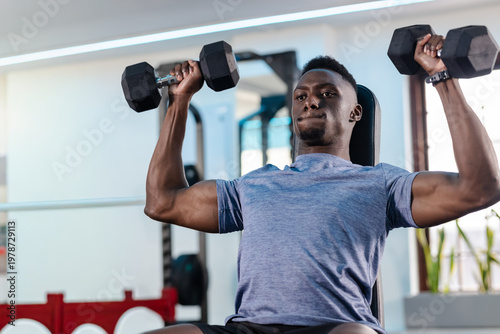 African American man in blue tee pressing hex dumbbells on adjustable bench at gym