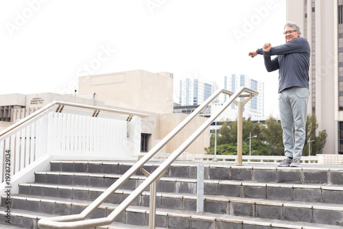 Mature man stretching arms across chest on plaza steps near metal rails in sportswear, copy space