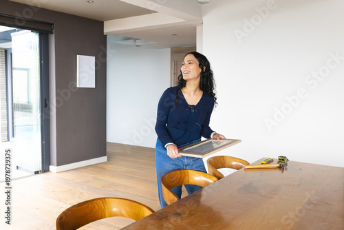 Woman in twenties standing in dining area beside table holding framed picture, using tape measure