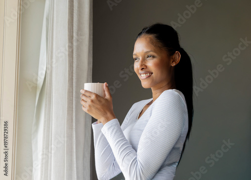 Photos Mid-adult Indian woman standing by window at home holding ceramic mug, wearing w