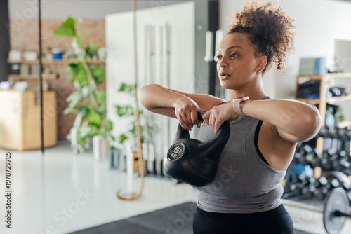Woman mid twenties lifting 5kg kettlebell on mat in fitness studio wearing gray top, copy space