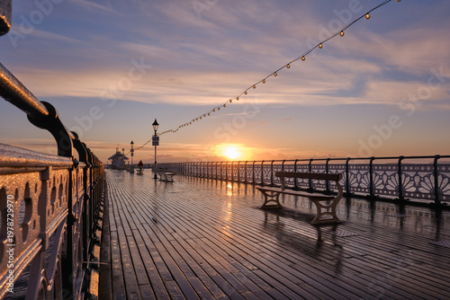 Sunrise blazes at the end of Penarth Pier, warm orange reflected in the wet decking as benches and ornate Victorian lamp posts line the rain-washed walkway.