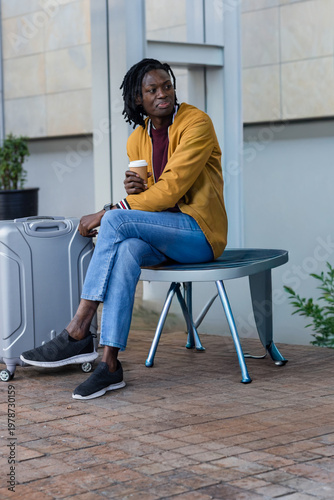 Mid-30s African American man sitting outside in yellow jacket holding coffee beside silver suitcase