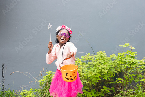 African American child wearing pink tutu and shutter shades holding pumpkin bucket and wand in yard