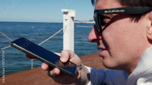 Man using voice assistant on smartphone at the beach, mobile technology, digital assistant, tech lifestyle, ocean view.