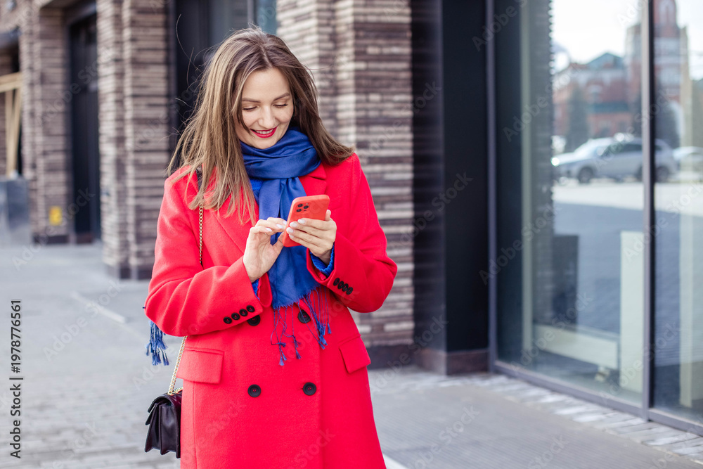 Fototapeta premium Young female tourist in red coat looking at smartphone while walking in modern city