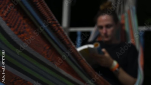 woman reading a book at night while gently swinging on a hammock on a house terrace. Soft lighting and calm atmosphere create a relaxing and intimate scene, ideal for concepts of leisure, solitude