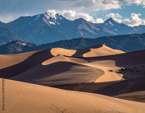 Vast dunes roll towards majestic snow-capped peaks under a partly cloudy sky. Shadows enhance the textures