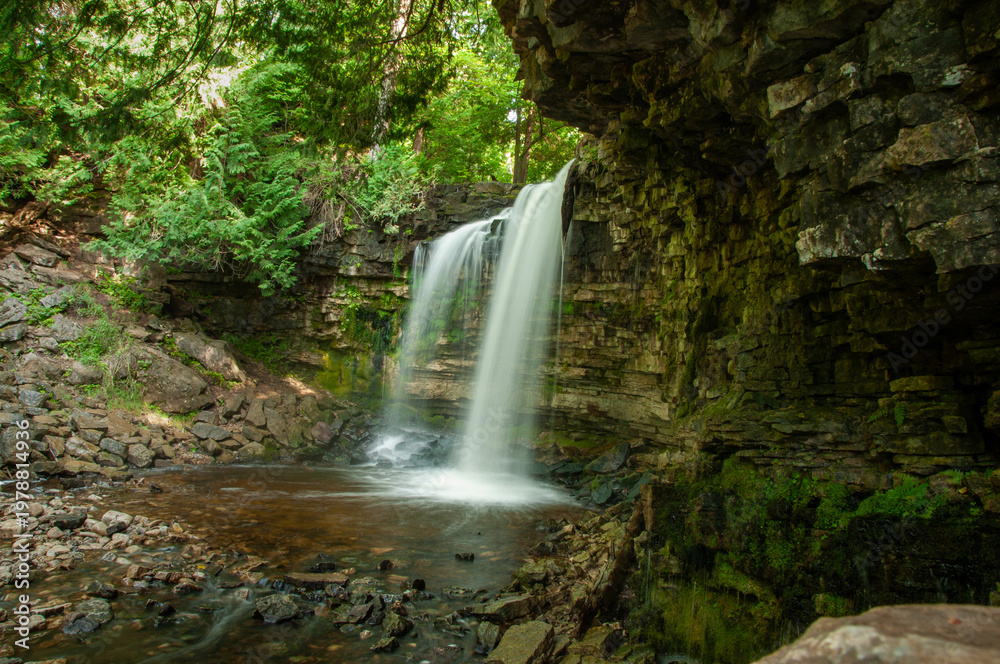 Obraz premium A long-exposure of a majestic waterfall cascading over a moss-covered cliff into a rocky pool. Surrounded by lush forest greenery and serene, peaceful natural scenery ideal for outdoor escape.