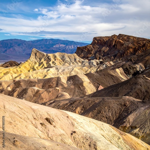 Vast landscape of colorful, textured hills bathed in sunlight. Mountains fade into a blue sky with soft, scattered clouds