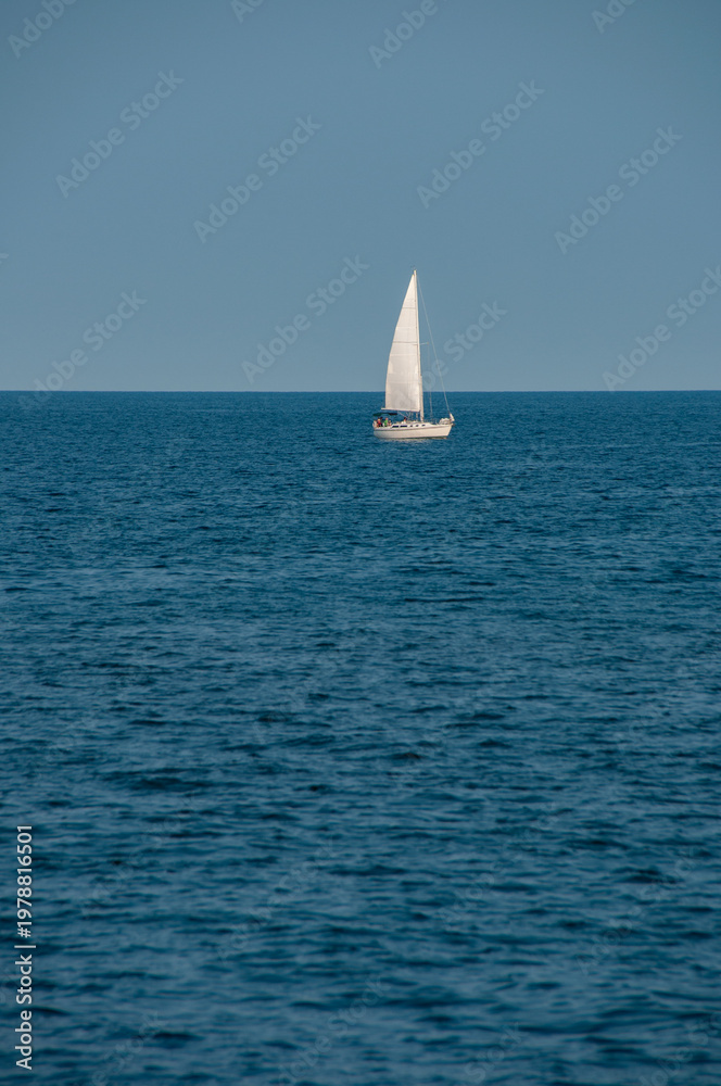 Fototapeta premium A lone white sailboat glides across a calm blue sea beneath a clear sky, highlighting open horizon, peaceful solitude and minimalist composition for a relaxed, nautical and leisurely summertime scene.