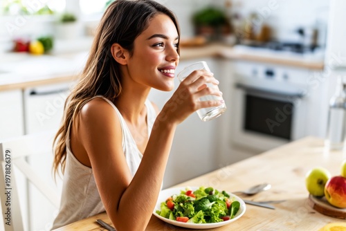 Woman Smiling While Eating Salad and Holding a Glass of Water in a Kitchen Se...