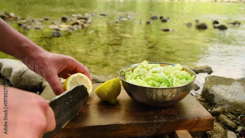 Close up of male hands cutting and squeeze lemon over fresh salad in bowl on wooden cutting board near wild river at summer time. Cooking outdoors and camping concept