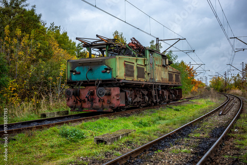 An old, green, electric shunting locomotive standing on a side track. The photo was taken on a cloudy autumn day.