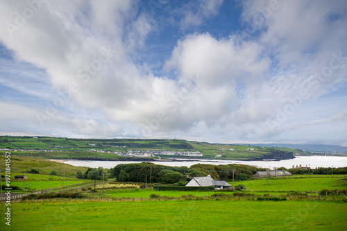 Lush green rolling hills and coastal village along Northern Ireland coastline under dramatic cloudy blue sky with calm bay waters and rural farmhouses in summer