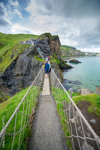 Tourists crossing Carrick-a-Rede Rope Bridge County Antrim Northern Ireland over turquoise coastal waters with dramatic cliffs and lush green grass under moody overcast skies on a summer day