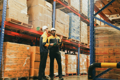 Two male employees work warehouse their duties including checking inventory moving goods forklift receiving shipping goods checking stock loading goods on pallets recording data into digital device.