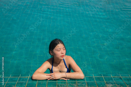 asian young woman in bathing suit relaxing on edge swimming pool in summer sunlight