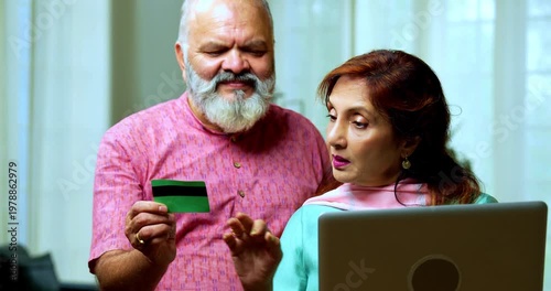Senior Indian couple standing in a modern living room, using a debit card for an online transaction and showing it to the camera with confidence and smiles