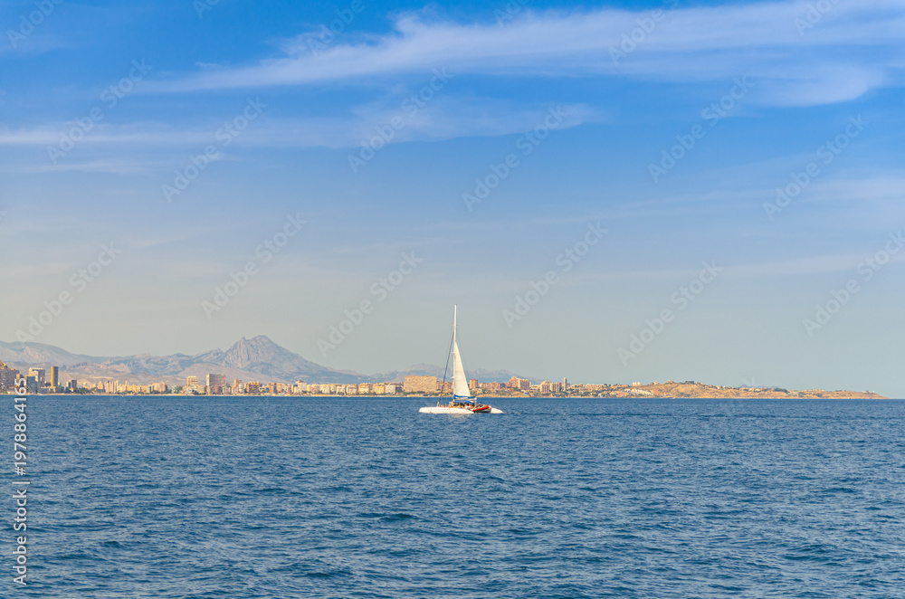 Fototapeta premium Catamaran with sail in water of Bahia Alicante bay Mediterranean Sea and Costa Blanca White coast, Cabo de la Huerta cape in Alicante city, mountain range background, Valencian Community, Spain