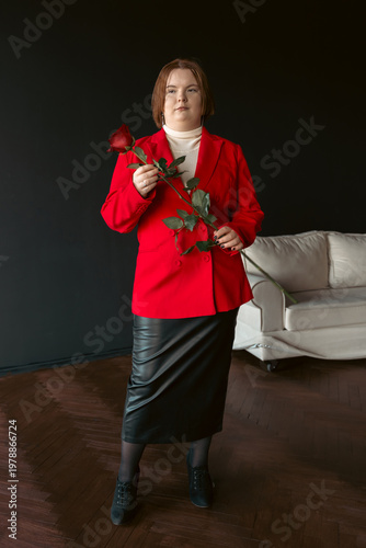 Full length portrait of a confident, stylish plus size young woman. She wears a vibrant red blazer, black leather skirt, and holds a single red rose, looking at the camera with a poised expression