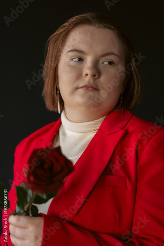 Studio portrait of a pensive young woman with short hair wearing a stylish red blazer and holding a rose. She has a thoughtful expression, looking away. Soft lighting on a dark background