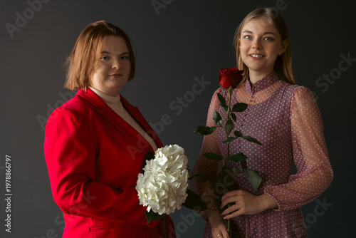 Portrait of two diverse friends. A woman with Down syndrome in a red blazer holds a white flower, her smiling friend holds a red rose. Soft studio light on a gray background. Inclusion concept