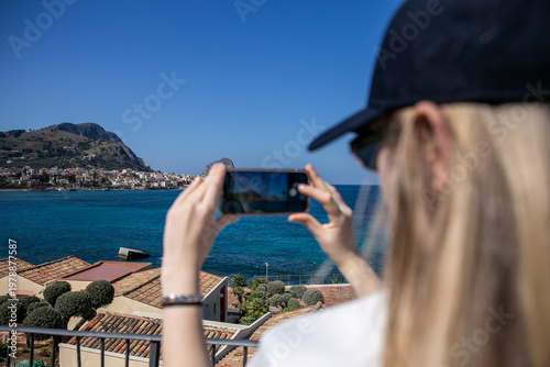 Tourist taking a photo with a smartphone at Domina Zagarella Sicily, enjoying a scenic coastal viewpoint with rocky shorelines and clear Mediterranean waters, capturing travel, leisure, and luxury