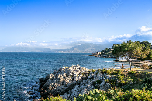 Scenic Mediterranean coastline near Palermo featuring rugged rocky formations, waves along the shore, and coastal vegetation with distant mountains under a clear blue sky, capturing the natural beauty