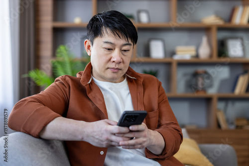 Asian man sitting comfortably on a sofa, looking intently at his smartphone, connecting, communicating, and interacting with technology in his modern home environment