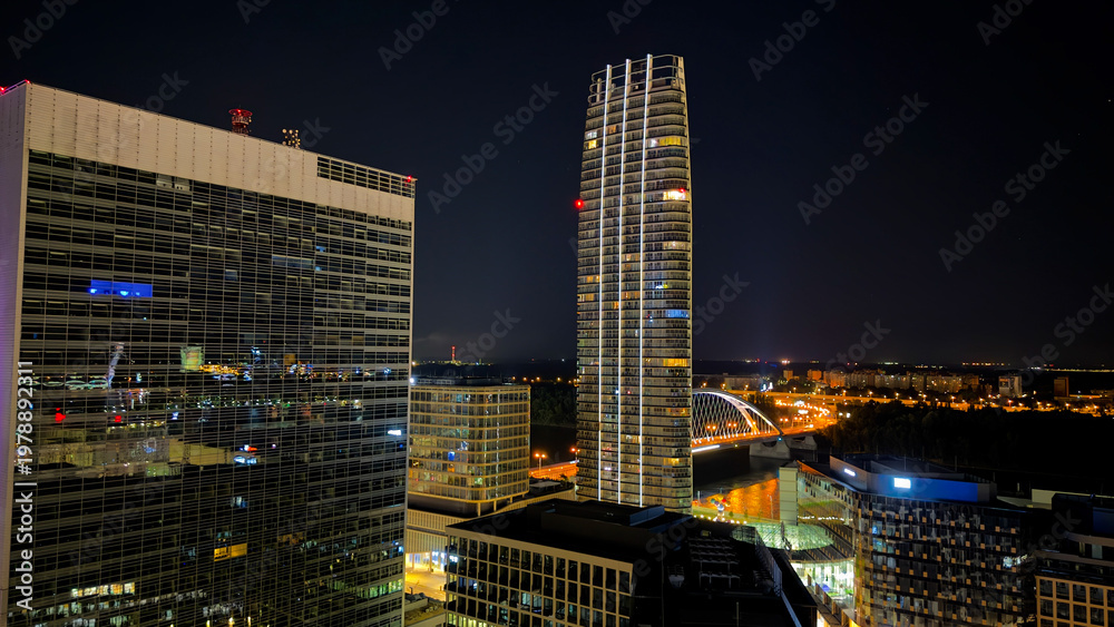 Fototapeta premium Modern city buildings with glass facade and tall skyscraper. Wide shot of office blocks reflecting night lights with a residential tower.