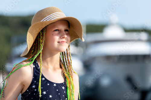 Girl wearing straw hat swimsuit stand sea luxury yachts marina during sunny summer vacation. Travel lifestyle, childhood freedom and seaside tourism atmosphere with blue water and resort background
