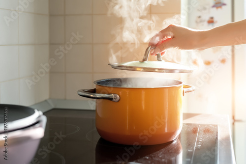 Slika na platnu Female hand open lid of enamel steel cooking pan on electric hob with boiling water or soup and scenic vapor steam backlit by warm sunlight at kitchen