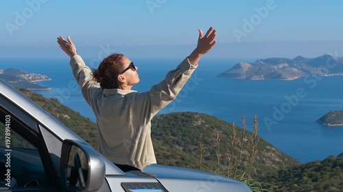 Woman raising arms in freedom admiring sea view. Happy woman on a road trip standing through the sunroof of a car with her arms outstretched, enjoying the breathtaking view of the sea with islands