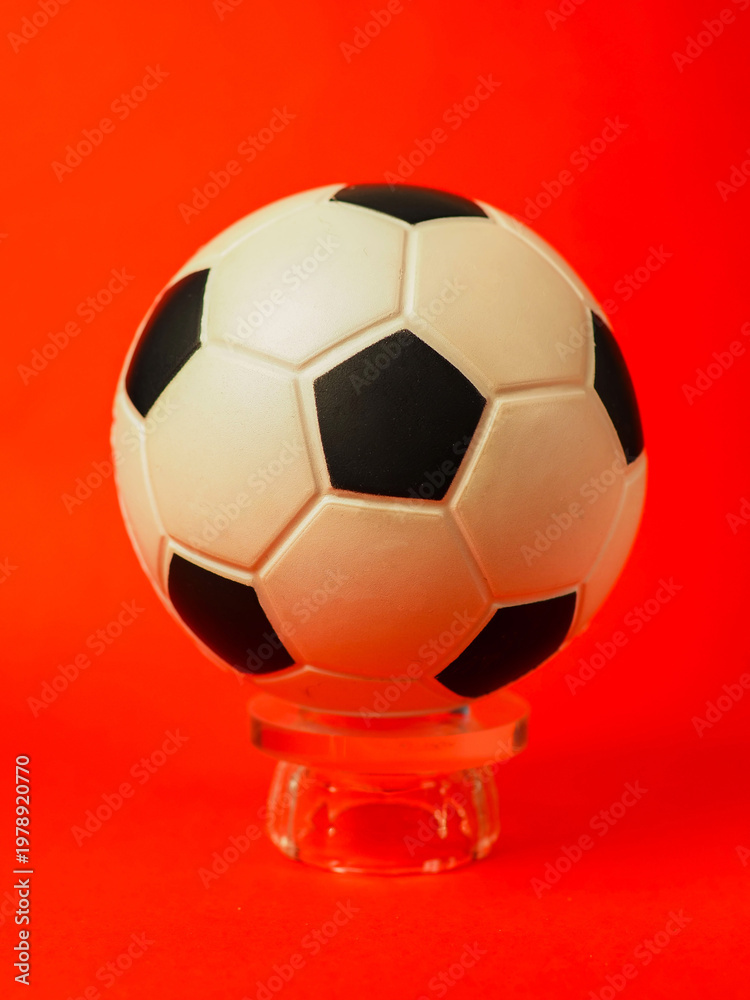 Naklejka premium Classic soccer ball rests on glass stand. Bright red background adds bold contrast. Surface texture shows subtle wear and shine. Clean composition ideal for sports or design use