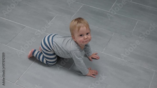 Caucasian infant explores the space indoors. A small blond child is on all fours. A joyful baby in striped pants and a gray sweater is getting ready to crawl around the house, smiling brightly