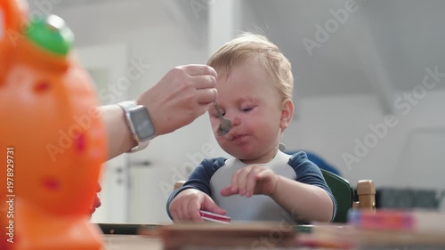 Caucasian baby spoon fed at table, parent hand feeding puree with spoon, orange toy blurred foreground, bib stained, baby tasting food, gentle caregiving in kitchen, soft natural light
