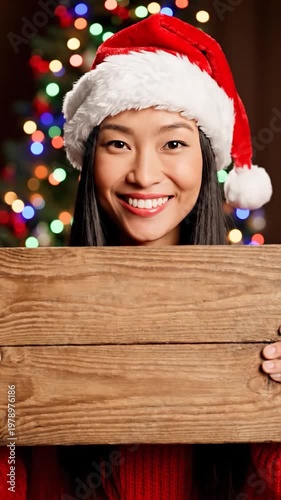 A smiling young woman wearing a festive red hat holds a blank wooden sign in front of a blurred holiday tree