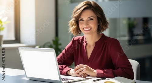 A smiling woman in a maroon blouse sits at a desk with a laptop, notepad, and pen in an office.