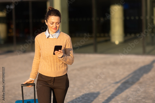 A woman in professional casual attire checks her mobile device for travel updates while walking with a rolling suitcase near a modern rail terminal at sunset.