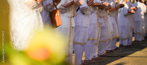 Obraz na płótnie Thai men in white walk in line with palms together carrying folded orange monk robes as medium to large group approaches temple doorway, warm light before ordination