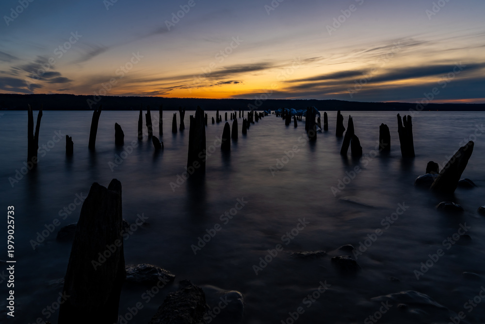 Obraz premium This tranquil landscape photo captures a serene shore at sunset. Old, weathered wooden pilings jut from calm waters, silhouetted against the bright sun and dark hills, evoking a peaceful mood.
